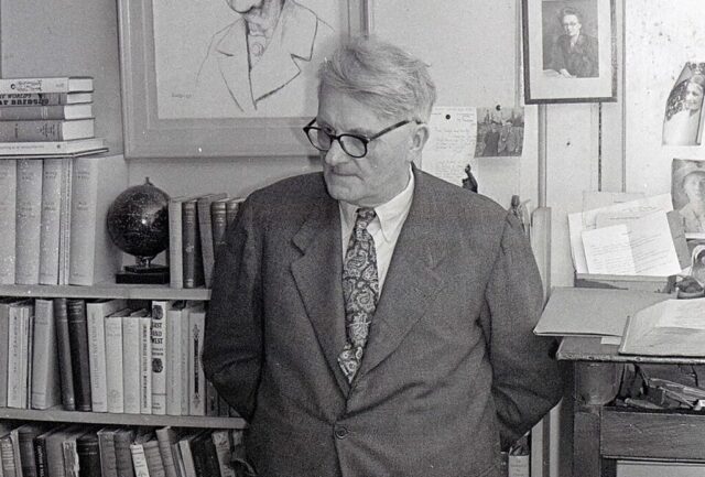 Black and white portrait of Joseph Needham in an office in Cambridge in 1965, looking down to the bottom left corner of the image.